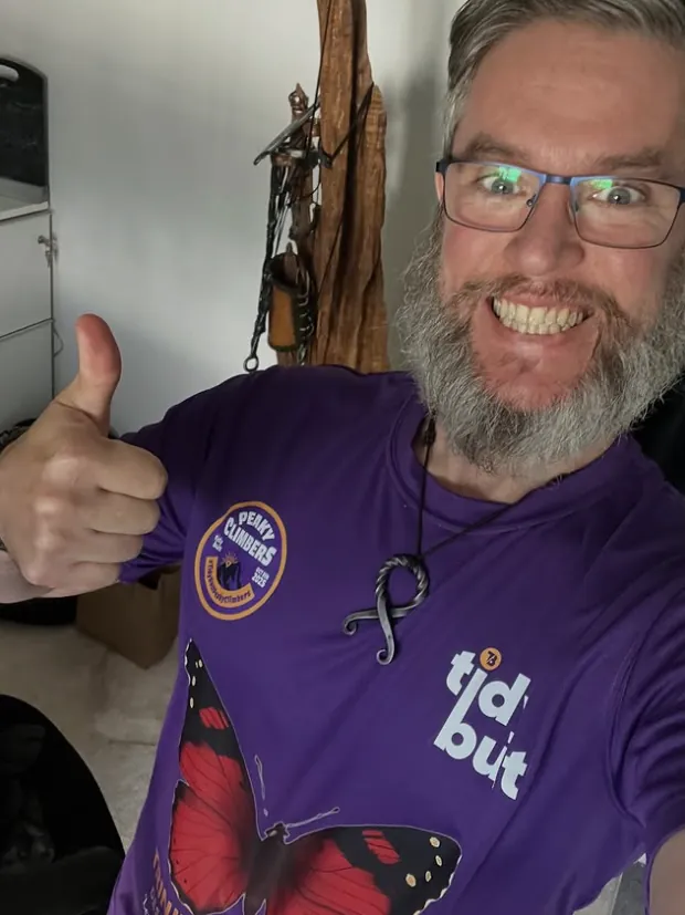 Wig grinning, thumbs up, in a Perky Climbers charity shirt with a forged pendant round his neck