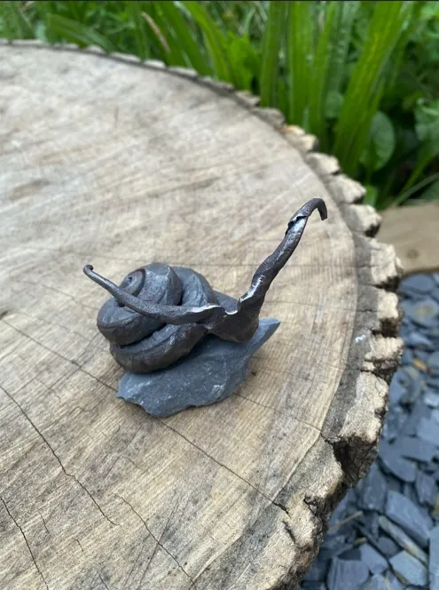 A snail forged from a scrap of rebar, sitting on a bit of slate on a log